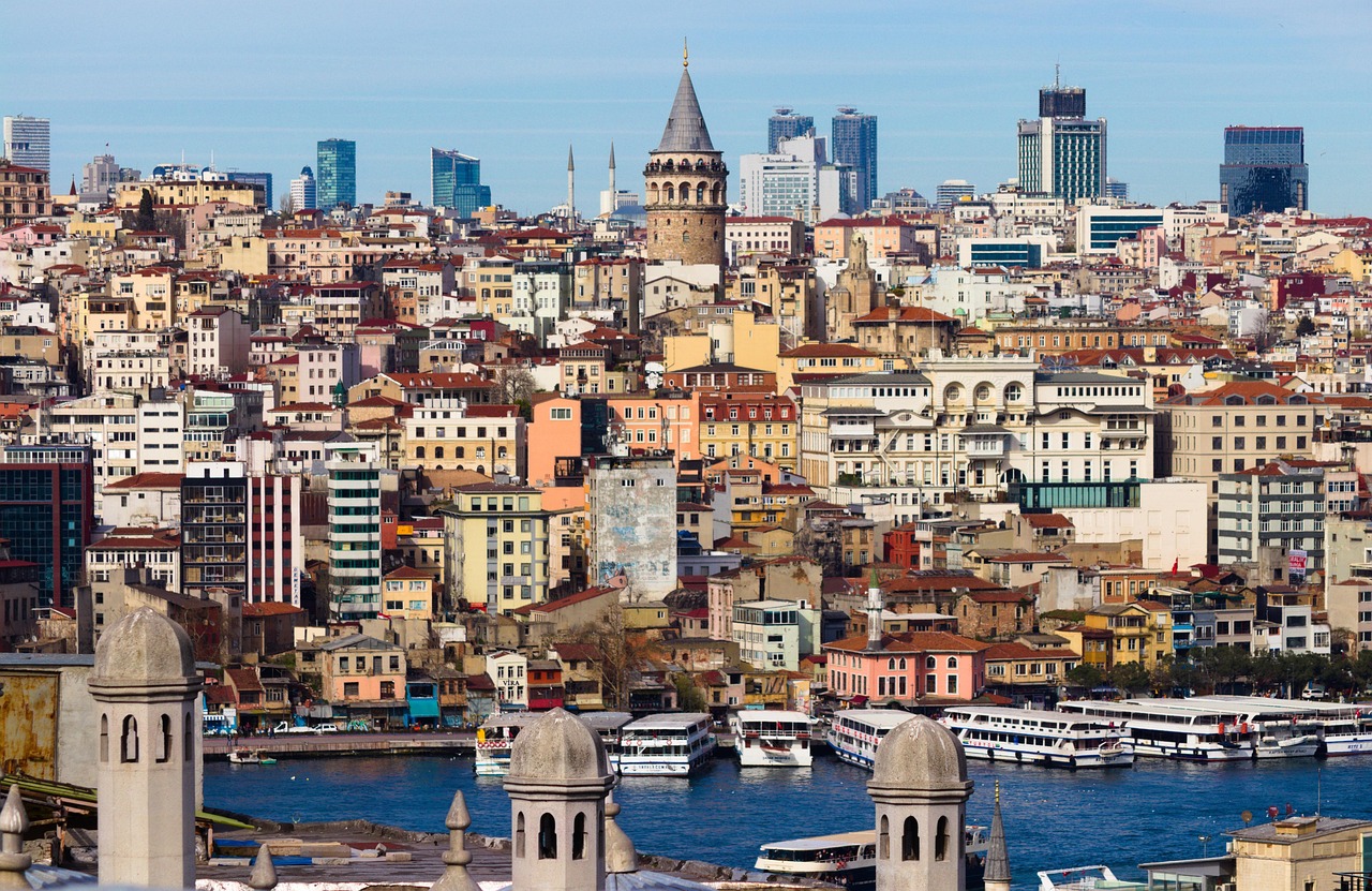 A bustling cityscape of Istanbul with the Galata Tower prominently visible, boats on the waterfront, and a mix of modern and historic architecture.