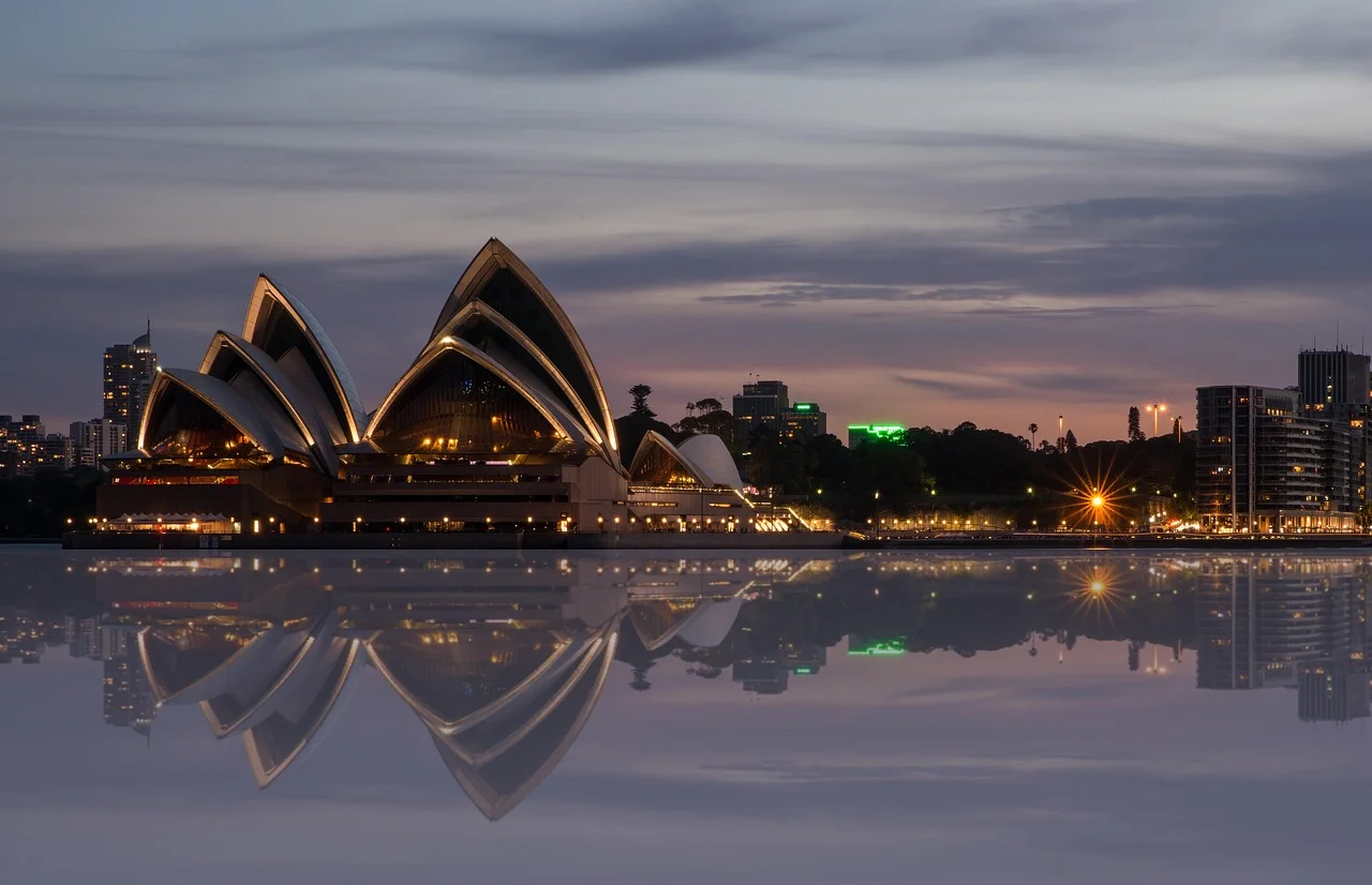 The Sydney Opera House illuminated at dusk, reflected in calm water with the city skyline in the background.