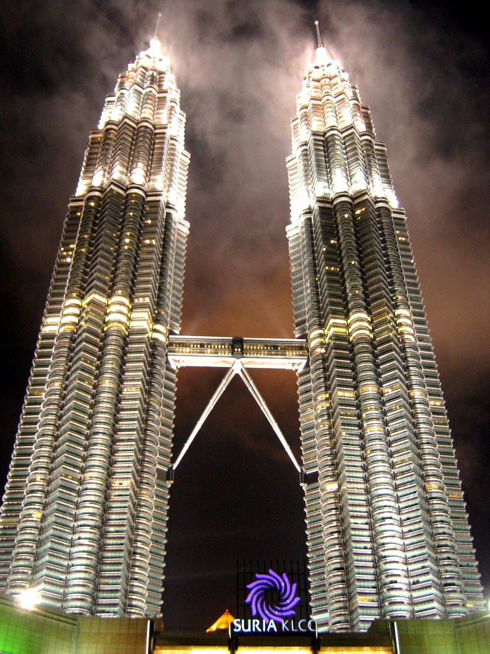 Two tall, illuminated twin towers connected by a skybridge at night.
