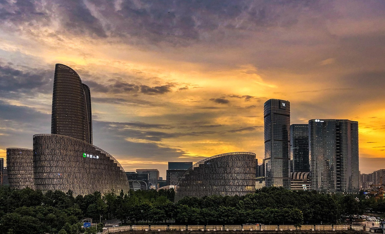 A city skyline at sunset with modern buildings and trees in the foreground.