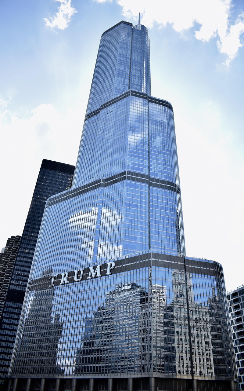 A tall skyscraper with the name Trump on it, reflecting other buildings in its glass facade.