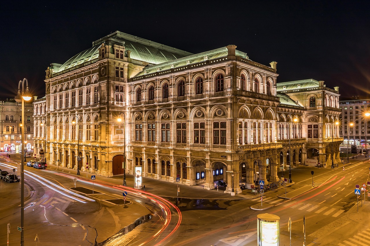 A grand, illuminated building with classical architecture at night, with light trails from passing cars on the street in front of it.