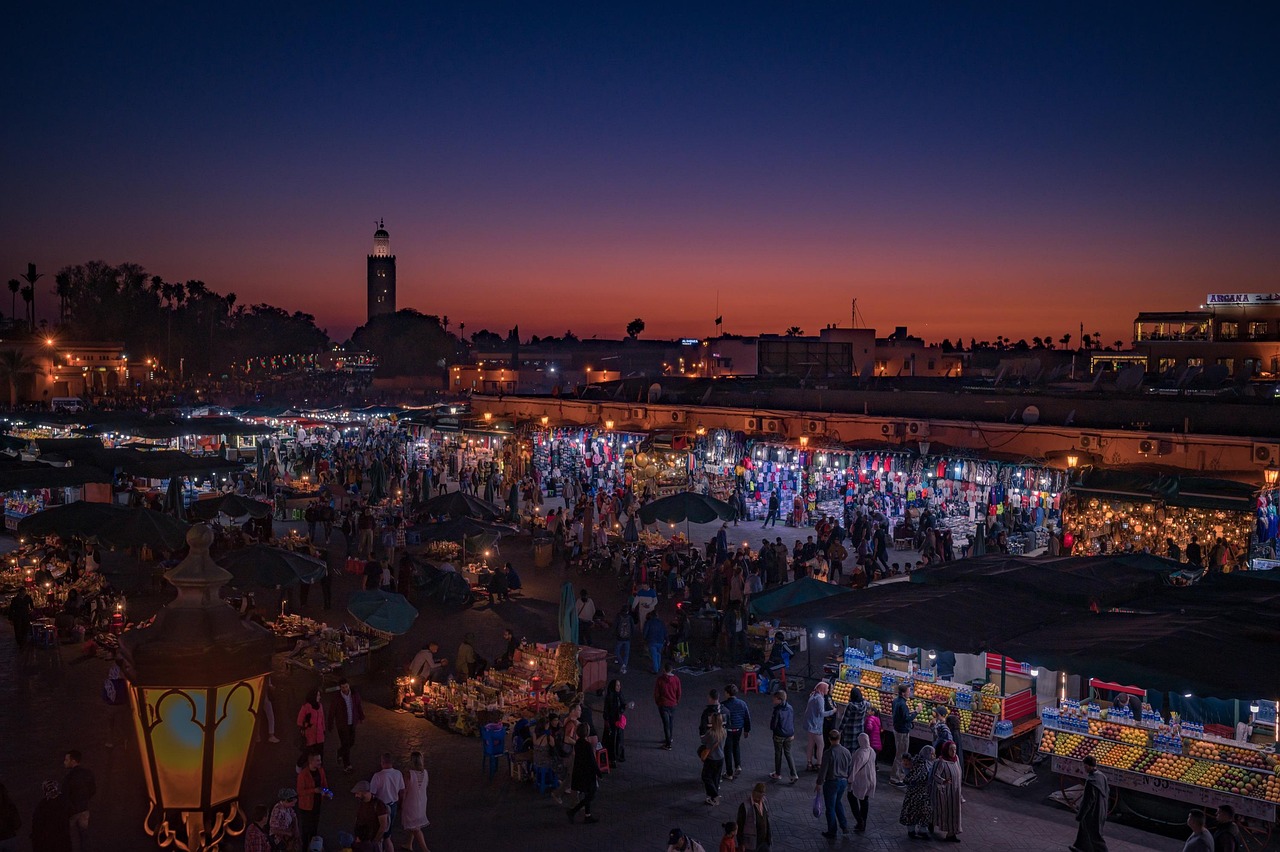 A bustling night market in Marrakech with numerous stalls and people shopping under colorful lights, with a prominent minaret in the background.