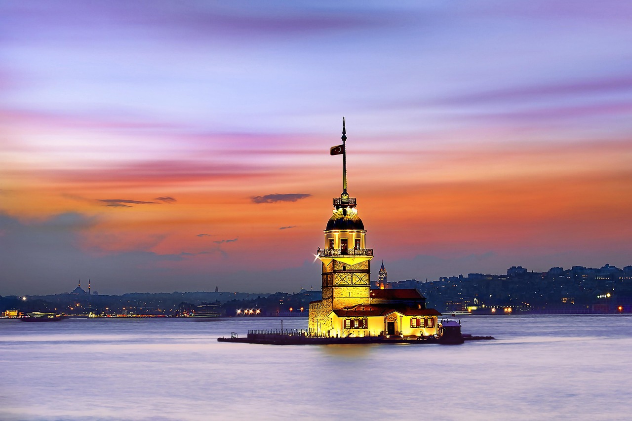 A tower illuminated at dusk, standing in the water with a city skyline in the background.