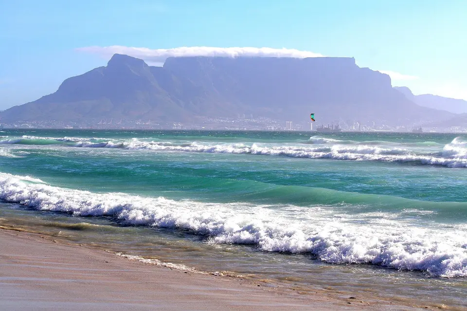 A scenic view of a beach with waves crashing onto the shore, a large mountain in the background, and a city skyline in the distance.