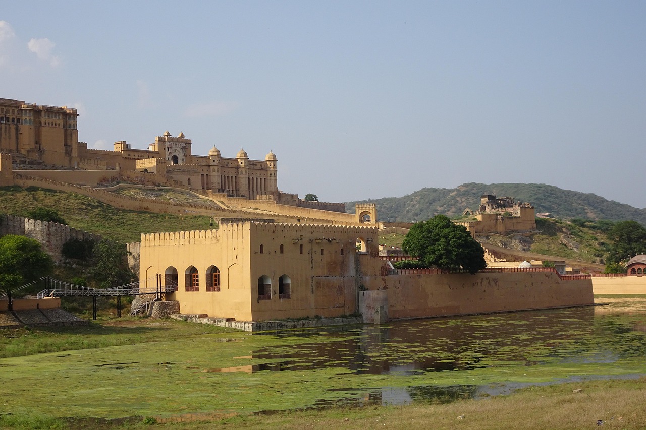 A large historical fort with yellowish-brown walls and multiple domed structures, situated on a hill with a water body in the foreground.