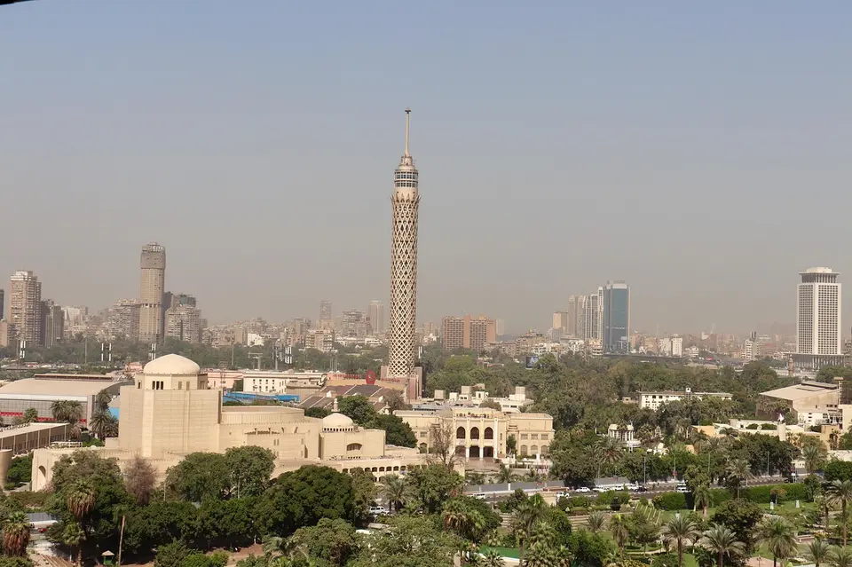 A cityscape featuring a tall skyscraper with a distinctive lattice design, surrounded by various buildings and greenery.