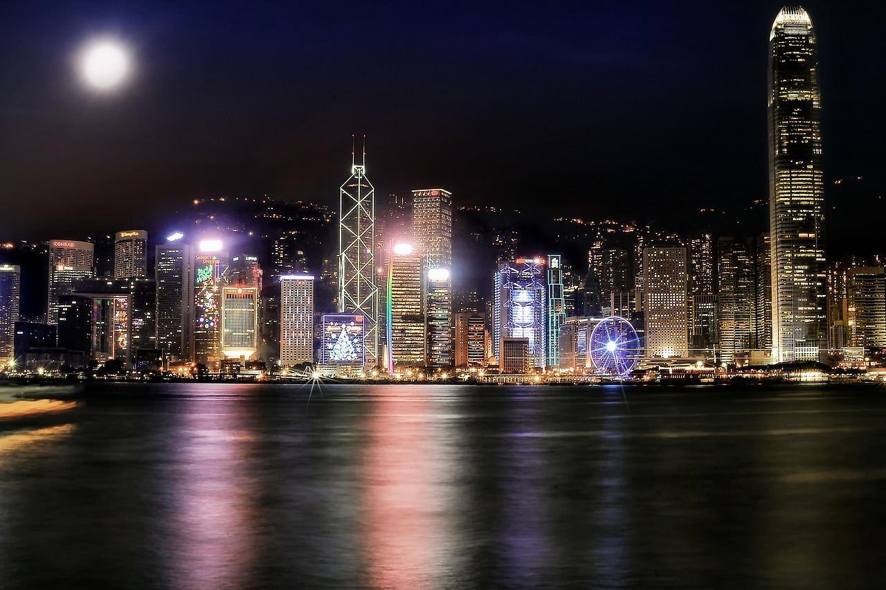 Nighttime cityscape of Hong Kong with illuminated skyscrapers and a Ferris wheel by the waterfront.
