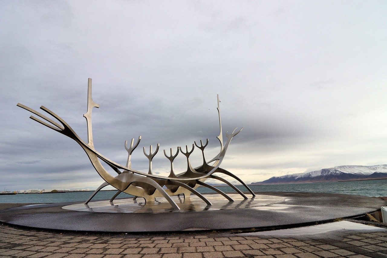 A large metallic sculpture of a Viking ship on a waterfront with mountains in the background.