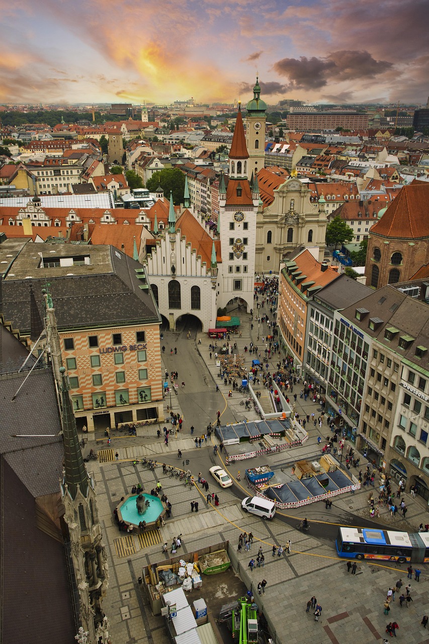 Aerial view of a bustling city square in Munich, Germany, featuring historic buildings, a clock tower, and a lively market with people and vehicles.