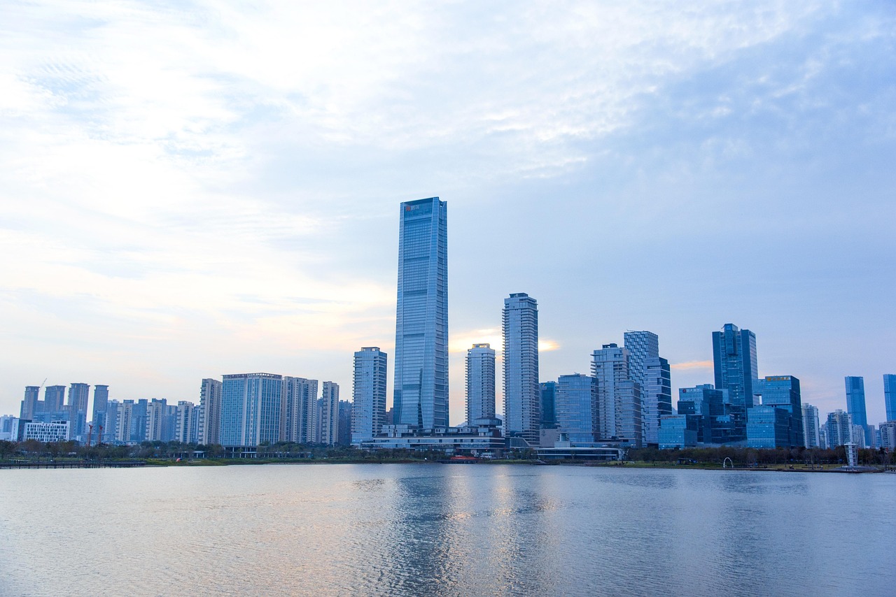 A city skyline with tall buildings reflected in a calm body of water during the day.