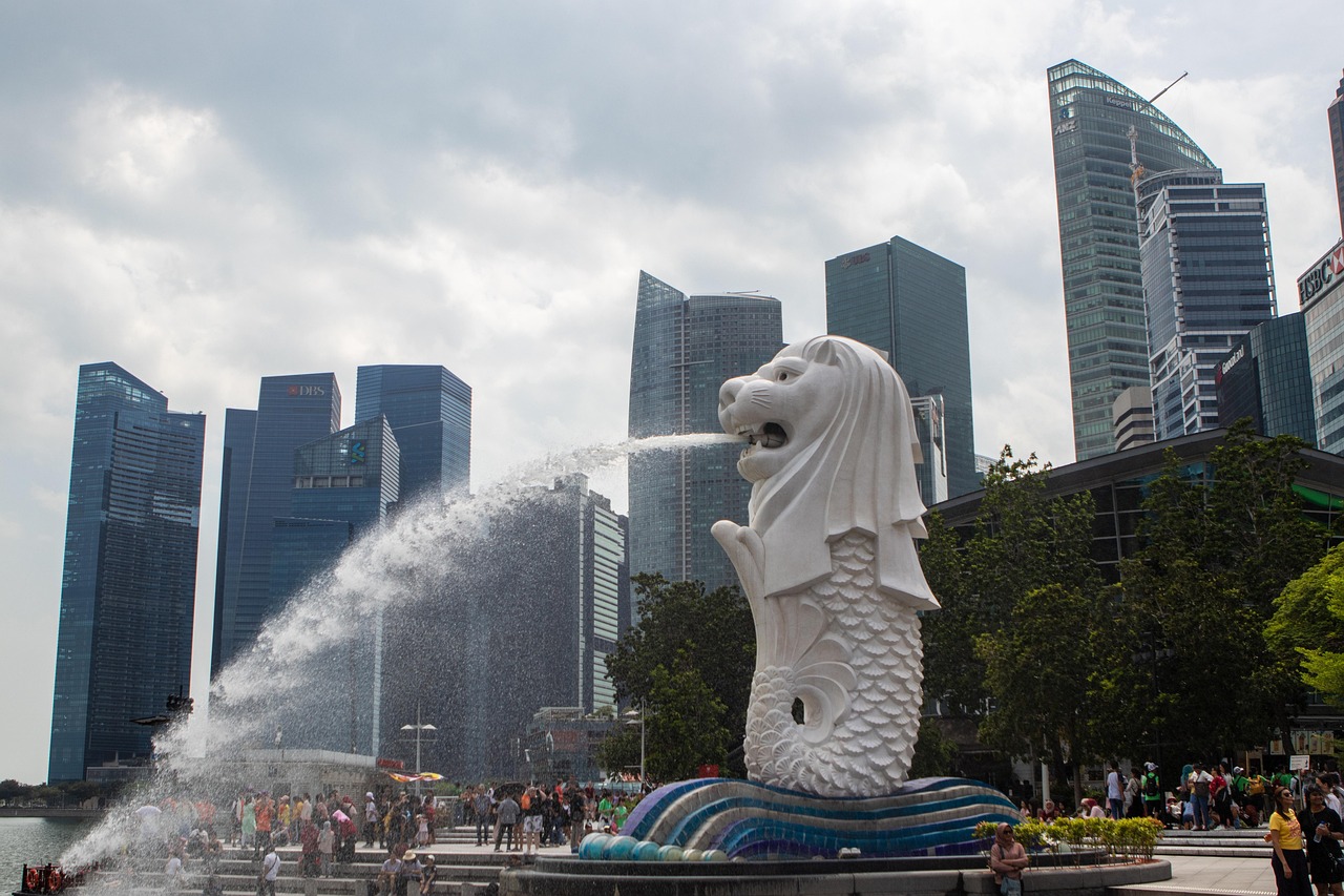 A large Merlion statue spouting water in front of a modern city skyline with tall buildings and a crowd of people in the foreground.