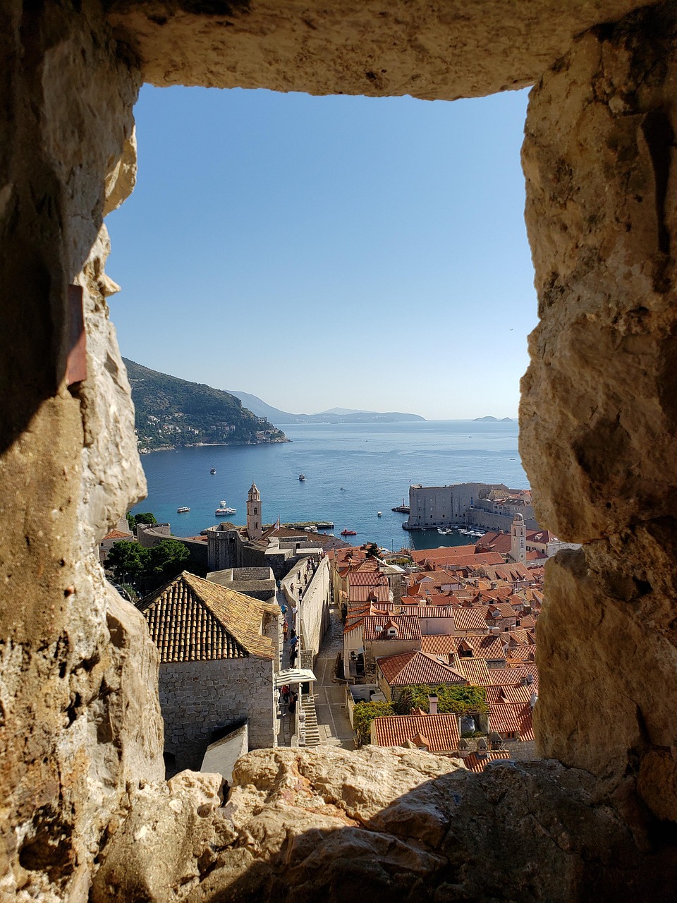 A view of a historic city with red-roofed buildings, a harbor, and a fortress in the distance, seen through an arched window.