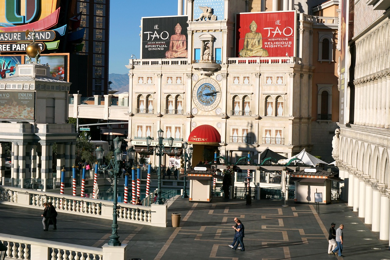 A bustling outdoor scene in front of a large, ornate building with a clock tower, featuring people walking and various signs for casinos and restaurants.