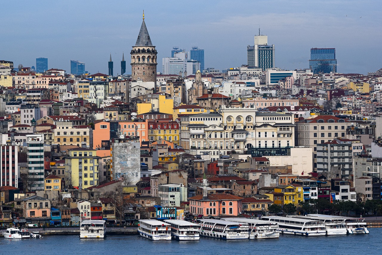 A bustling cityscape of Istanbul with the Galata Tower prominently featured, boats docked along the waterfront, and a mix of modern and historic buildings in the background.
