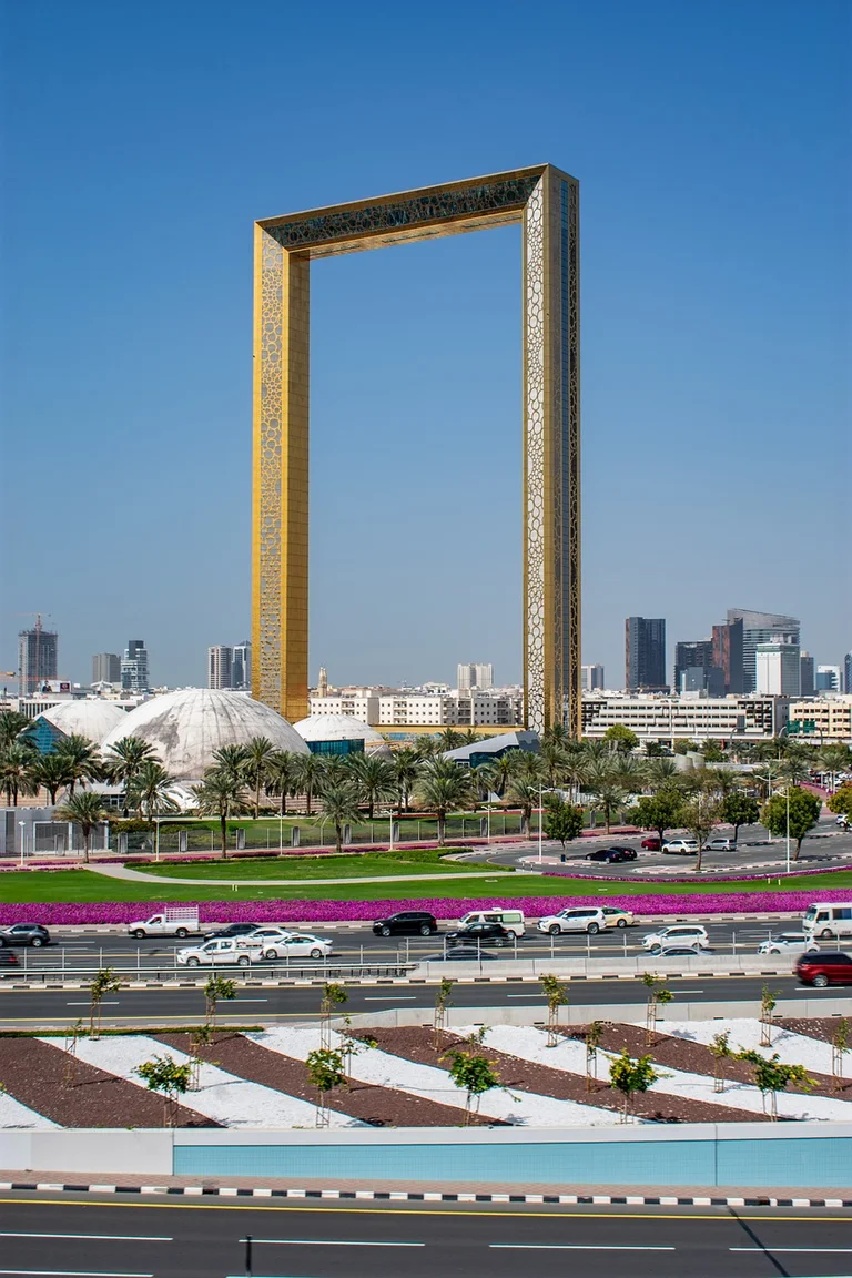 A large golden rectangular frame structure in a city with a modern skyline, palm trees, and busy roads.