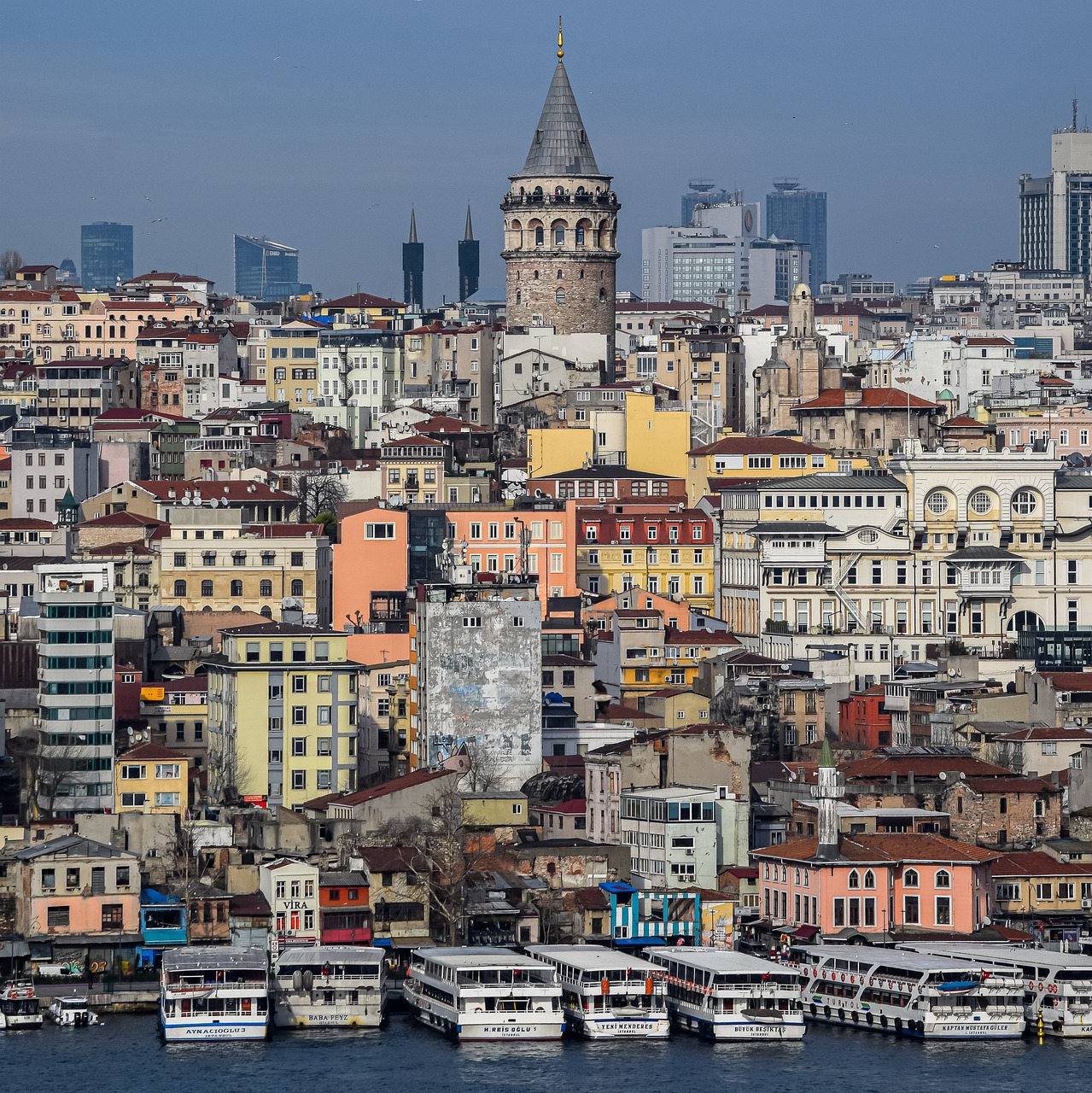 A bustling cityscape of Istanbul with the Galata Tower prominently featured, surrounded by colorful buildings and boats docked along the waterfront.