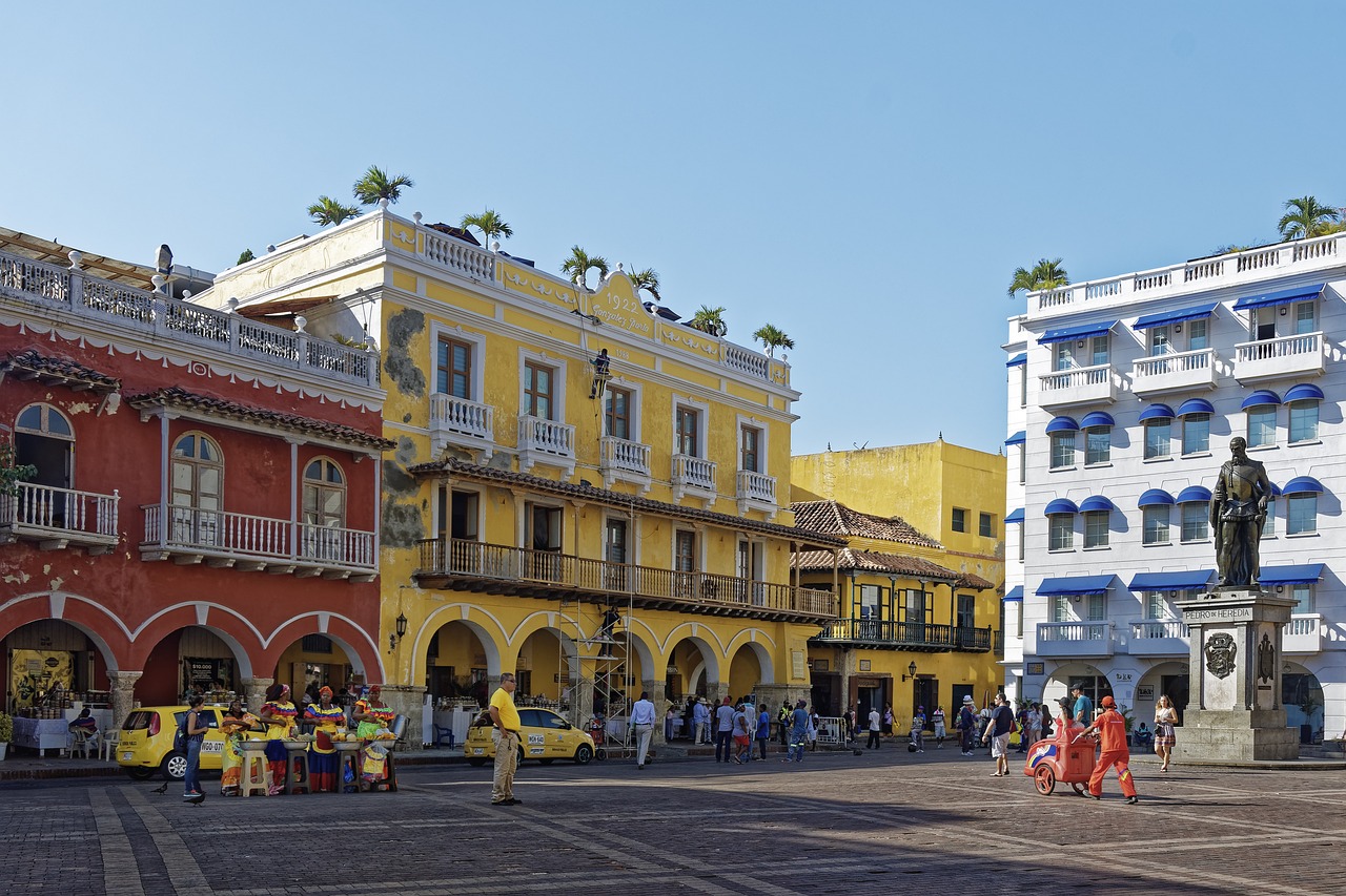 A bustling street scene in Cartagena, Colombia, with colorful colonial buildings, a statue, and people walking and shopping.