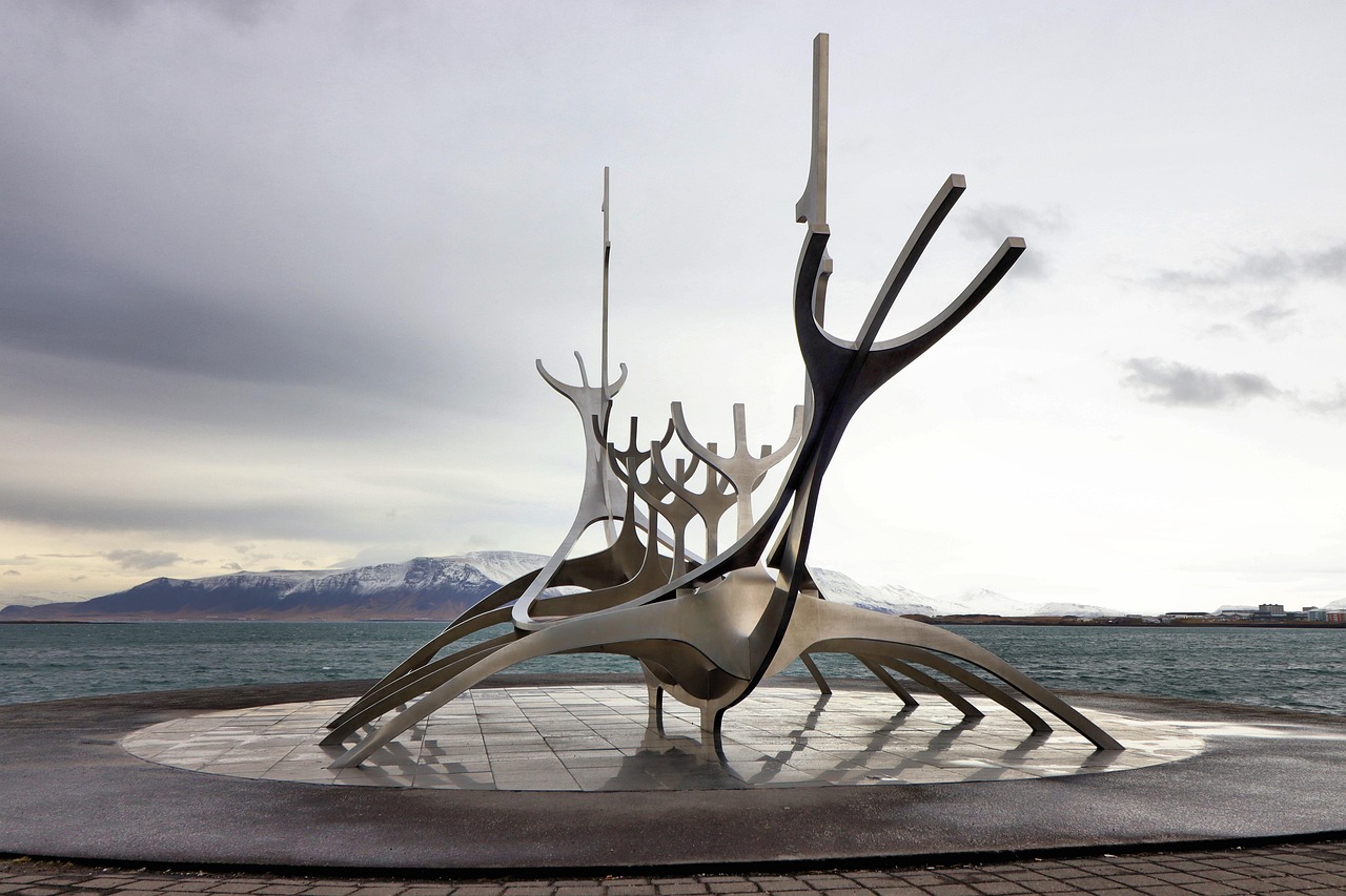 A large metallic sculpture resembling a Viking ship on a waterfront with mountains in the background.