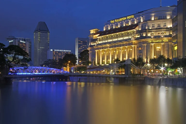 Night view of a city waterfront with illuminated buildings and a blue-lit bridge.