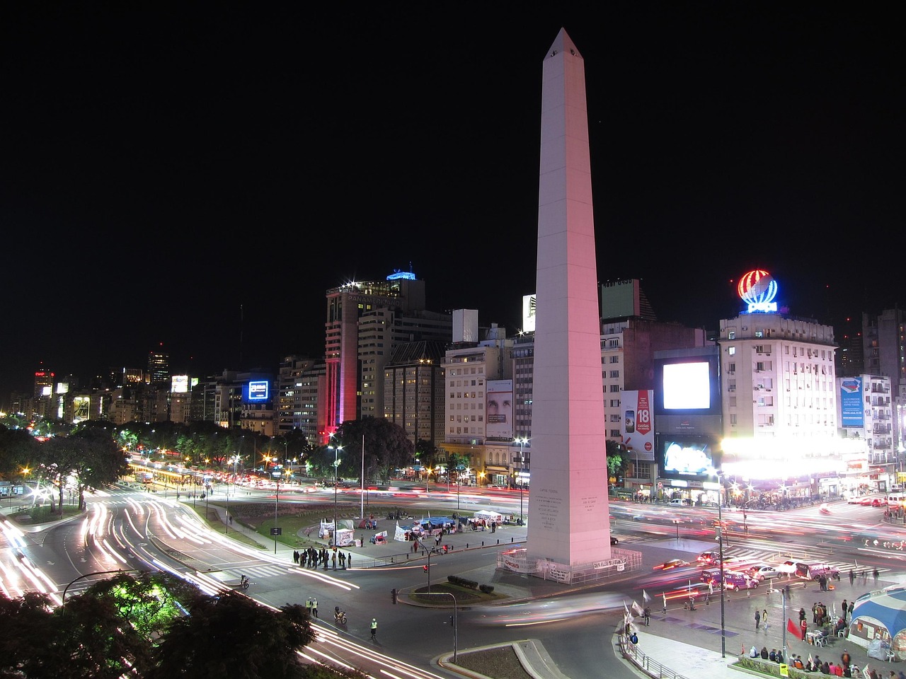 Night view of a bustling city with a tall obelisk monument illuminated in pink and blue lights, surrounded by traffic and buildings.