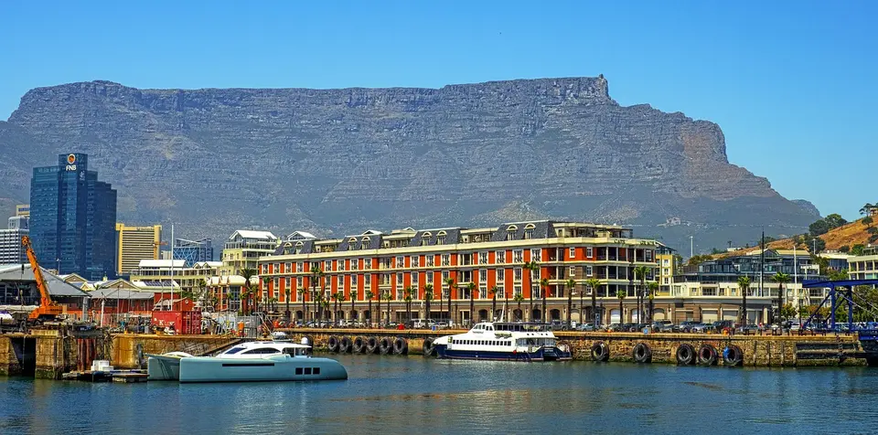 A waterfront cityscape with a large mountain in the background, boats docked in the foreground, and colorful buildings along the water.