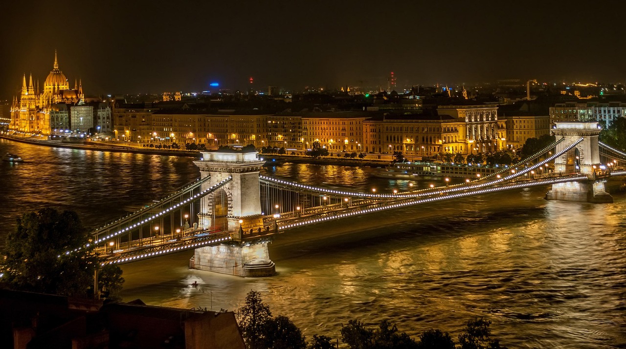Night view of a city with a lit bridge over a river, prominent buildings, and a domed structure in the background.