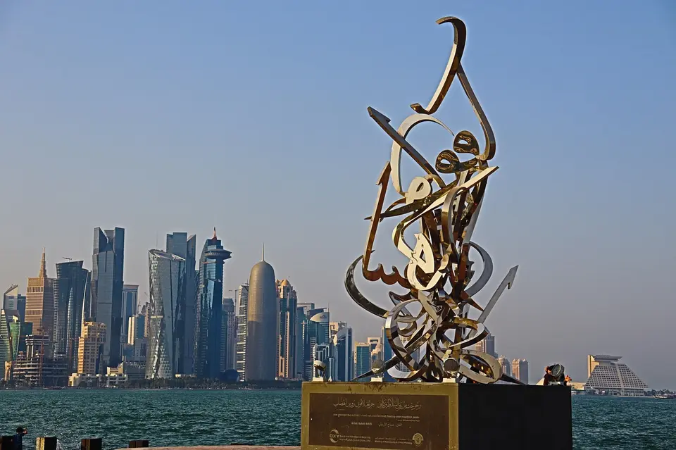 A modern city skyline with tall buildings and a metallic sculpture in the foreground, situated by the water.