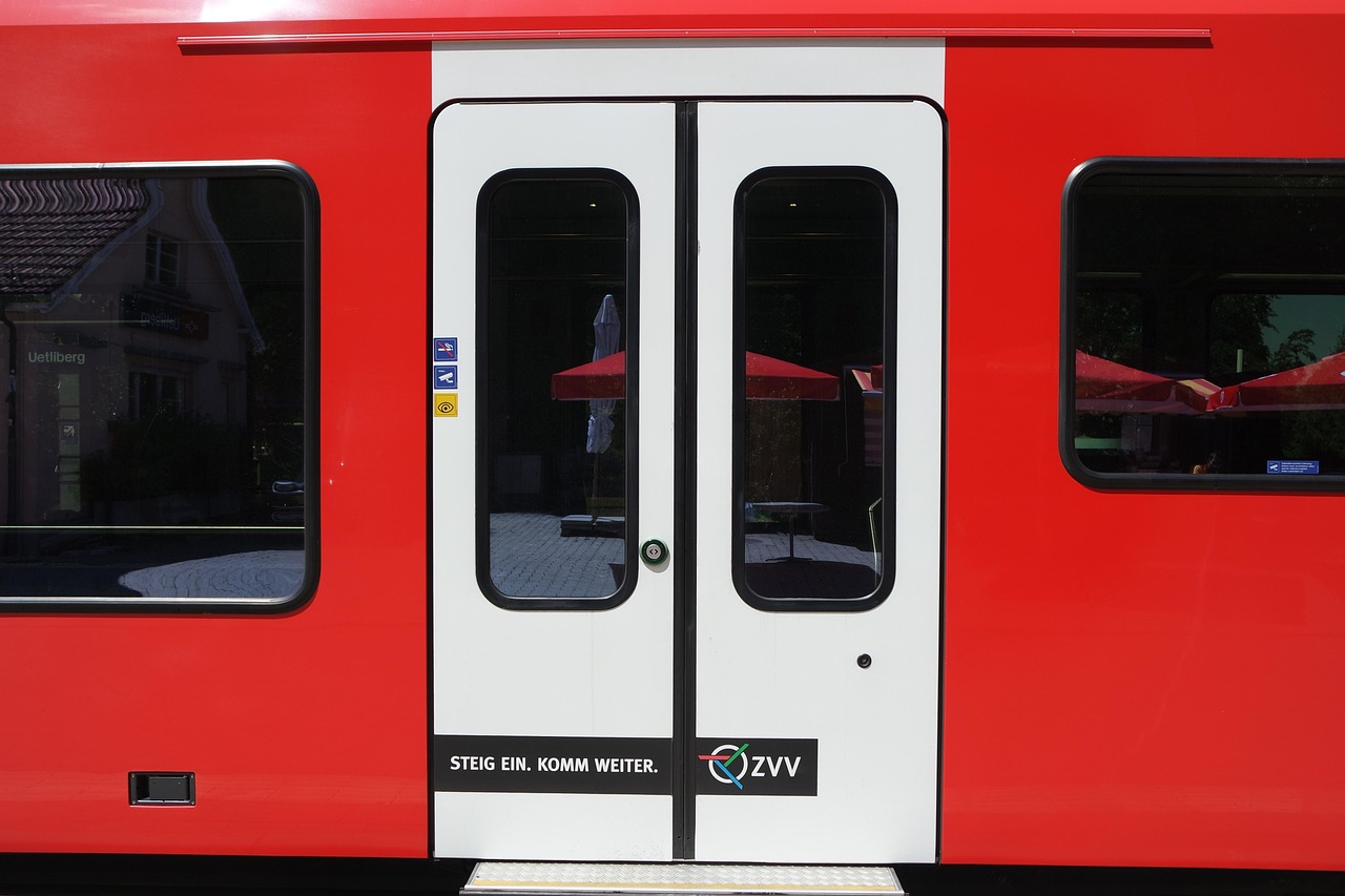 A red train with white doors, reflecting outdoor scenery, including red umbrellas and buildings.
