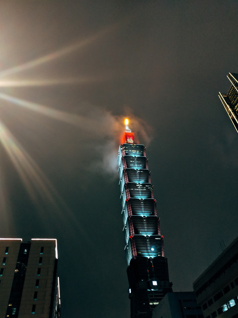 A tall skyscraper illuminated at night with a red and orange glow at the top, surrounded by other buildings.