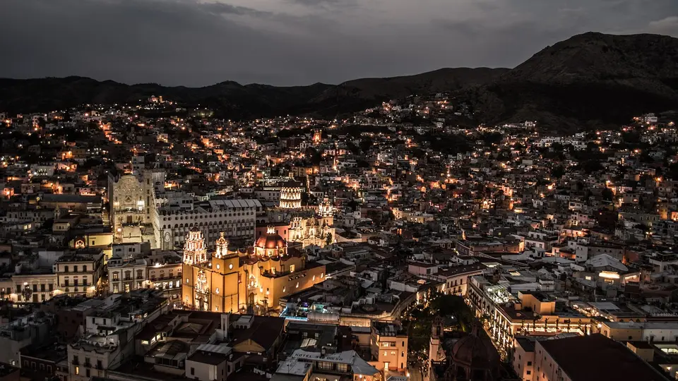 Nighttime view of a densely populated city with illuminated buildings and a prominent church in the foreground, surrounded by mountains.