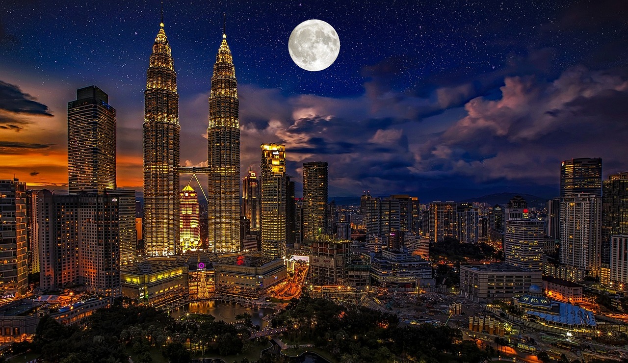 Nighttime cityscape of Kuala Lumpur with the Petronas Twin Towers prominently featured under a full moon.