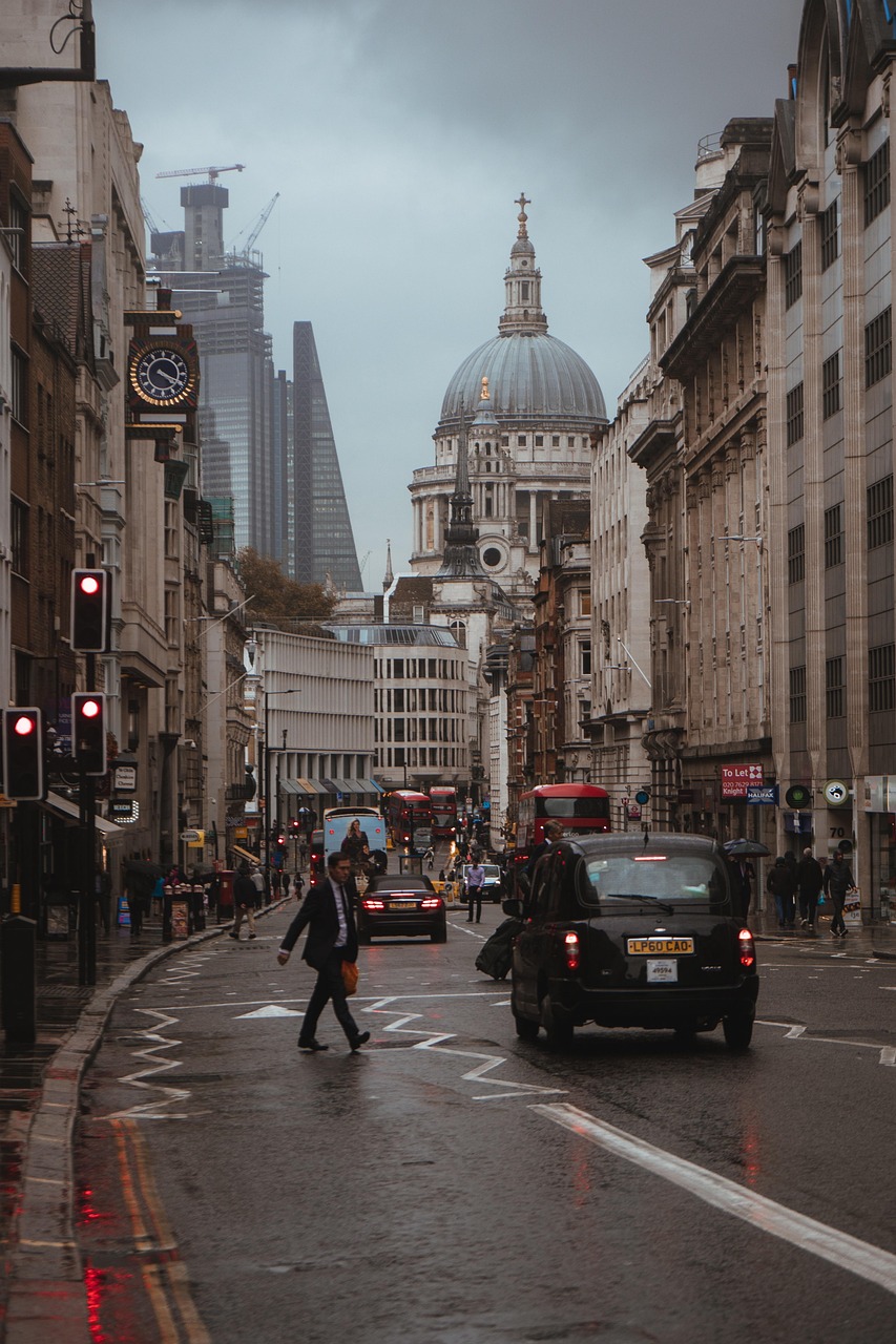 A bustling street in London with historic architecture, including St. Paul's Cathedral in the background, and modern skyscrapers. The street is wet, indicating recent rain, with pedestrians and vehicles navigating the area.