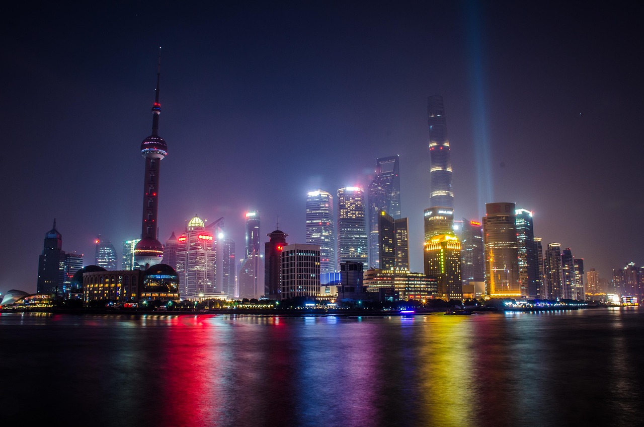 Night view of a city skyline with tall buildings and a tower, reflected in the water.