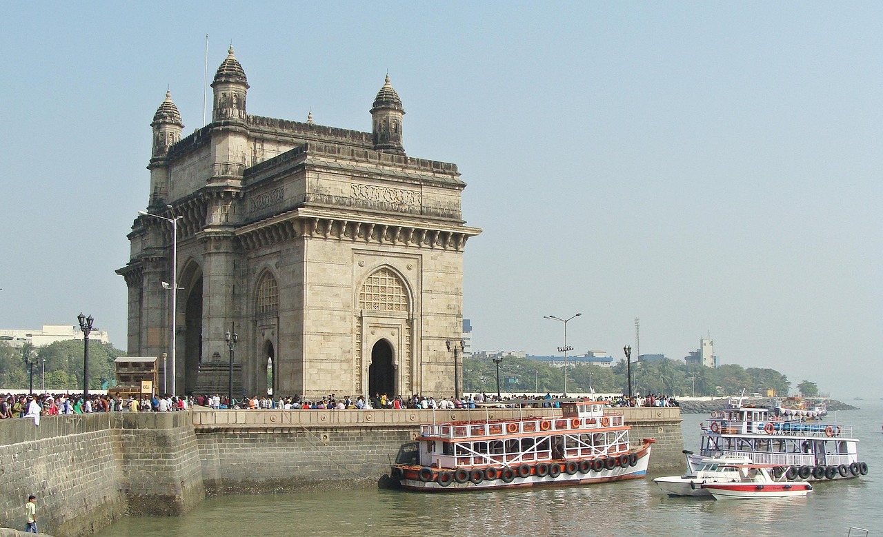 A large, historic archway known as the Gateway of India in Mumbai, India, with boats in the water and many people gathered around.