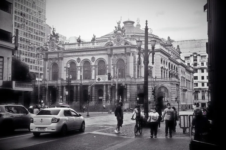 A black and white photograph of a bustling street in front of a grand theater building with ornate architecture, pedestrians, and vehicles.