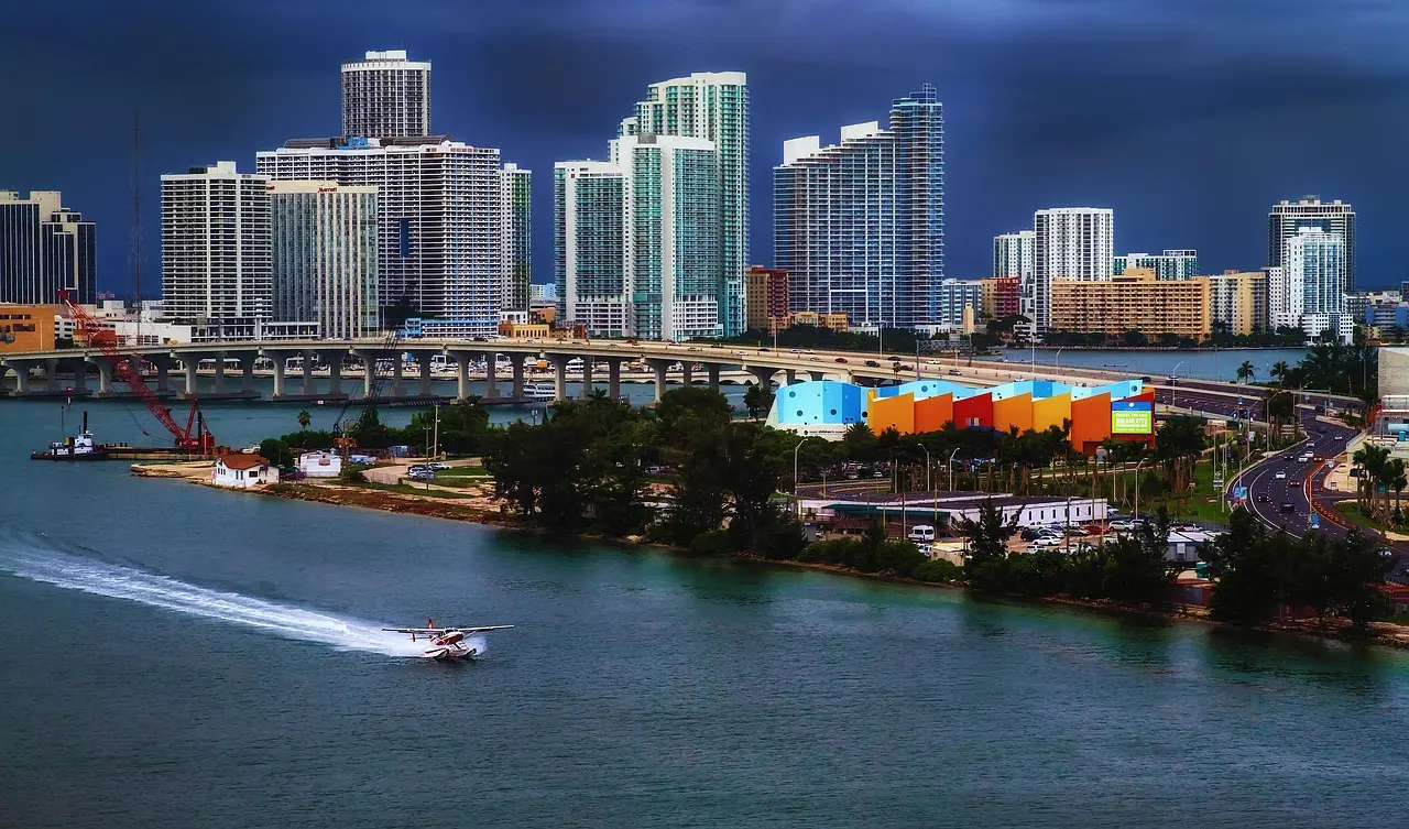 A city skyline with tall buildings, a bridge, and a highway over water at dusk. A boat is seen on the water in the foreground.