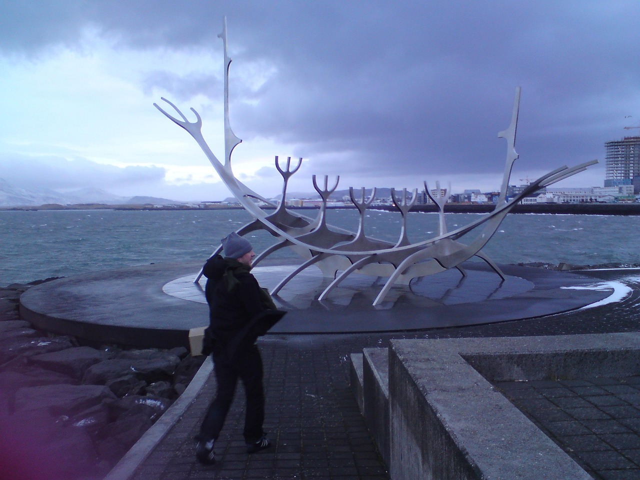 A person standing near a large, metallic Viking ship sculpture by the waterfront in Reykjavik, Iceland.