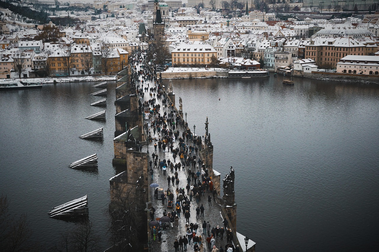 Aerial view of a historic bridge covered in snow with people walking across it, surrounded by a cityscape with snow-covered buildings and a river on either side.
