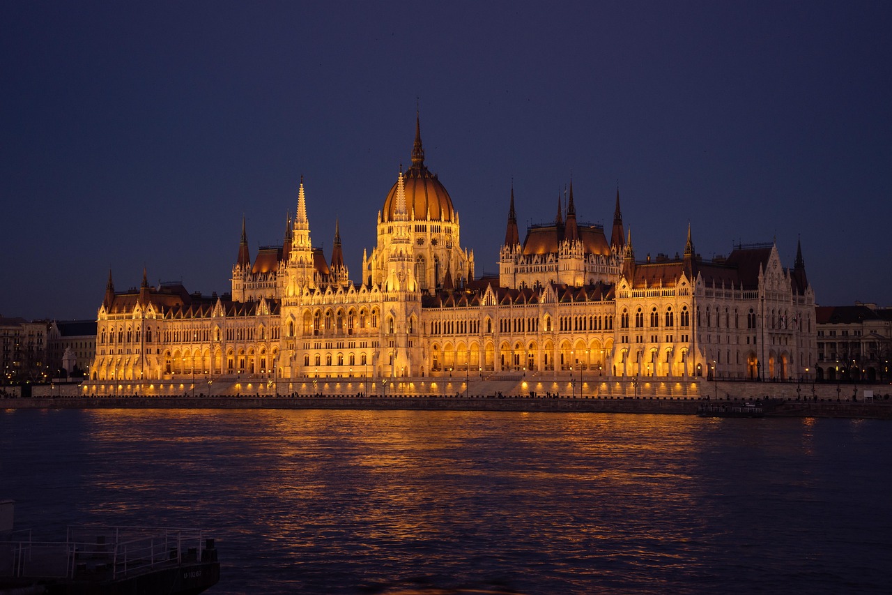A grand, illuminated building with a large dome and multiple spires reflected in a body of water at night.