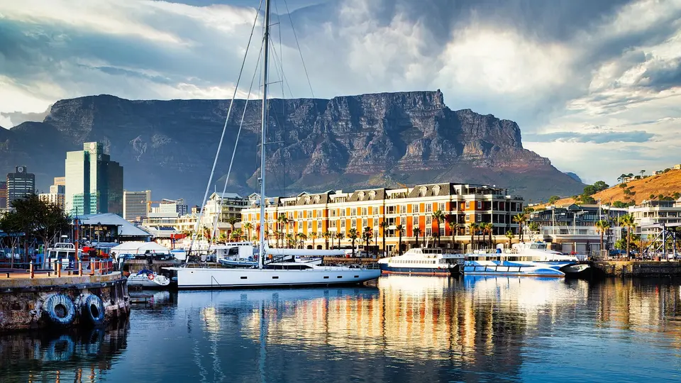 A scenic view of a waterfront city with boats docked, colorful buildings, and a large mountain in the background.