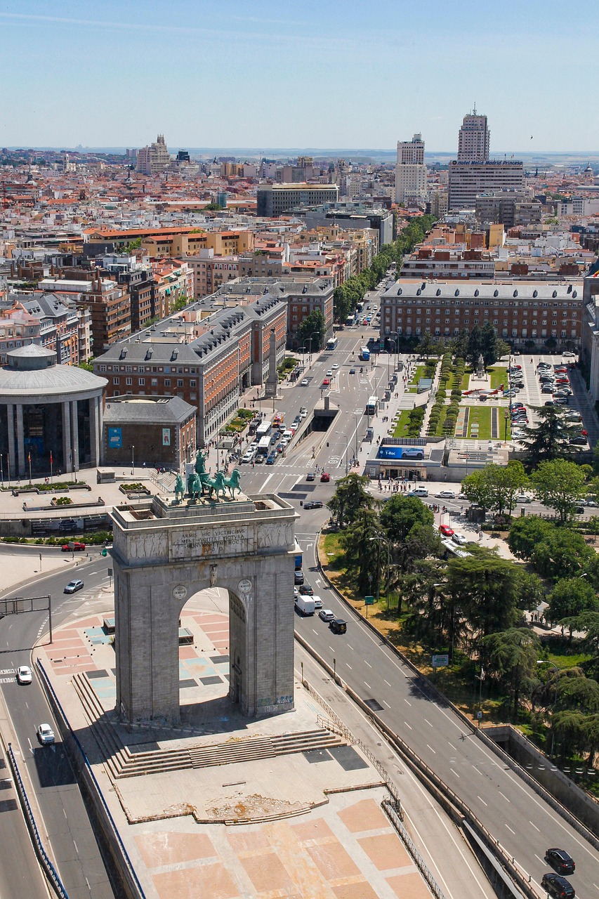 Aerial view of a city with a prominent arch monument in the foreground, wide streets, and various buildings in the background.