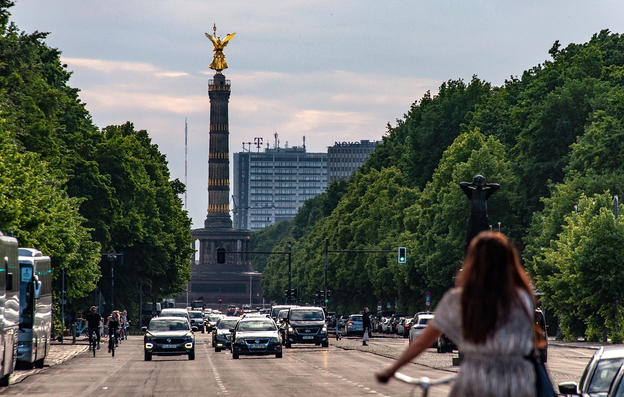 A bustling city street with a tall column monument in the background, surrounded by trees and buildings.