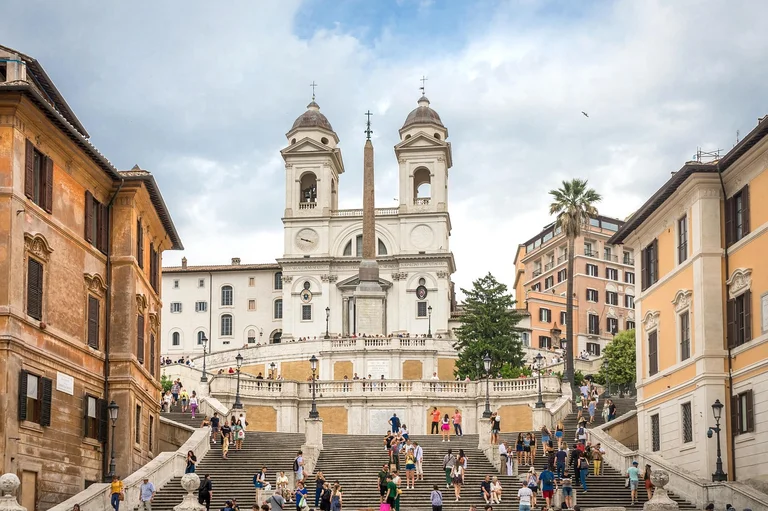 A bustling scene at the Spanish Steps in Rome, Italy, with people ascending and descending the stairs, surrounded by historic buildings and a clear sky.