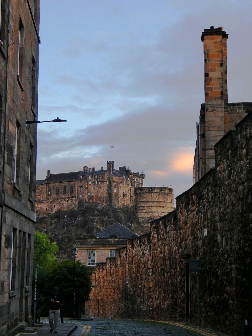 A person walking down a cobblestone street in Edinburgh with Edinburgh Castle visible in the background at dusk.