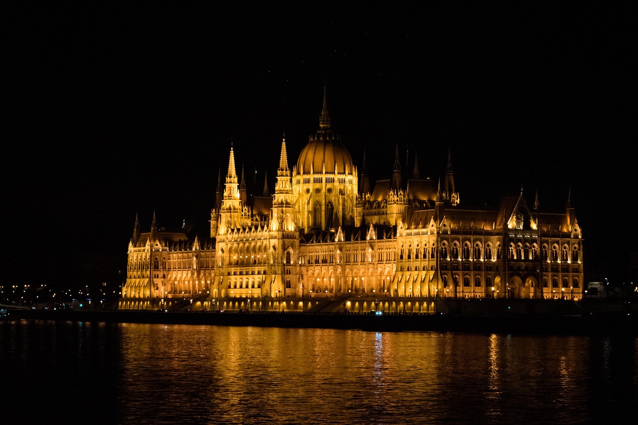 A grand, illuminated building with a large dome and multiple spires reflected in a body of water at night.