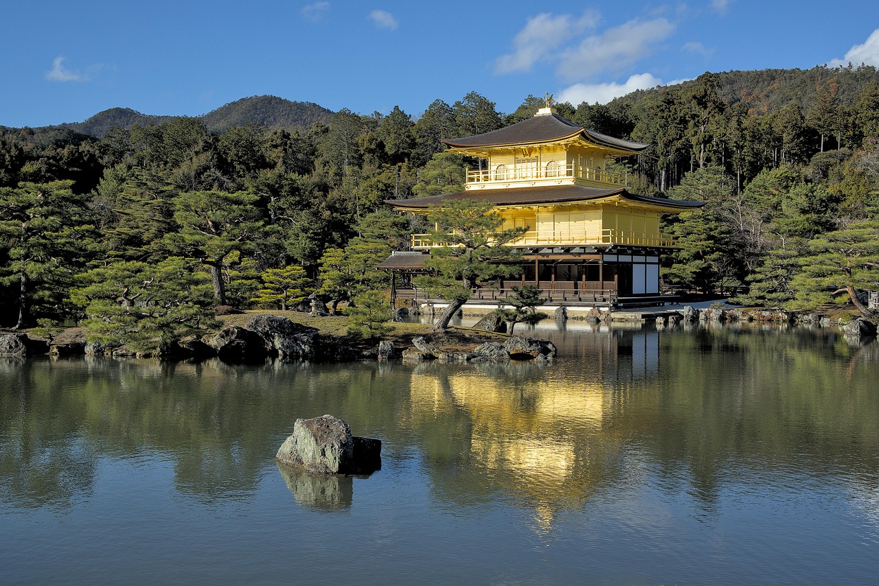 A golden pavilion reflected in a calm pond surrounded by lush greenery and mountains in the background.
