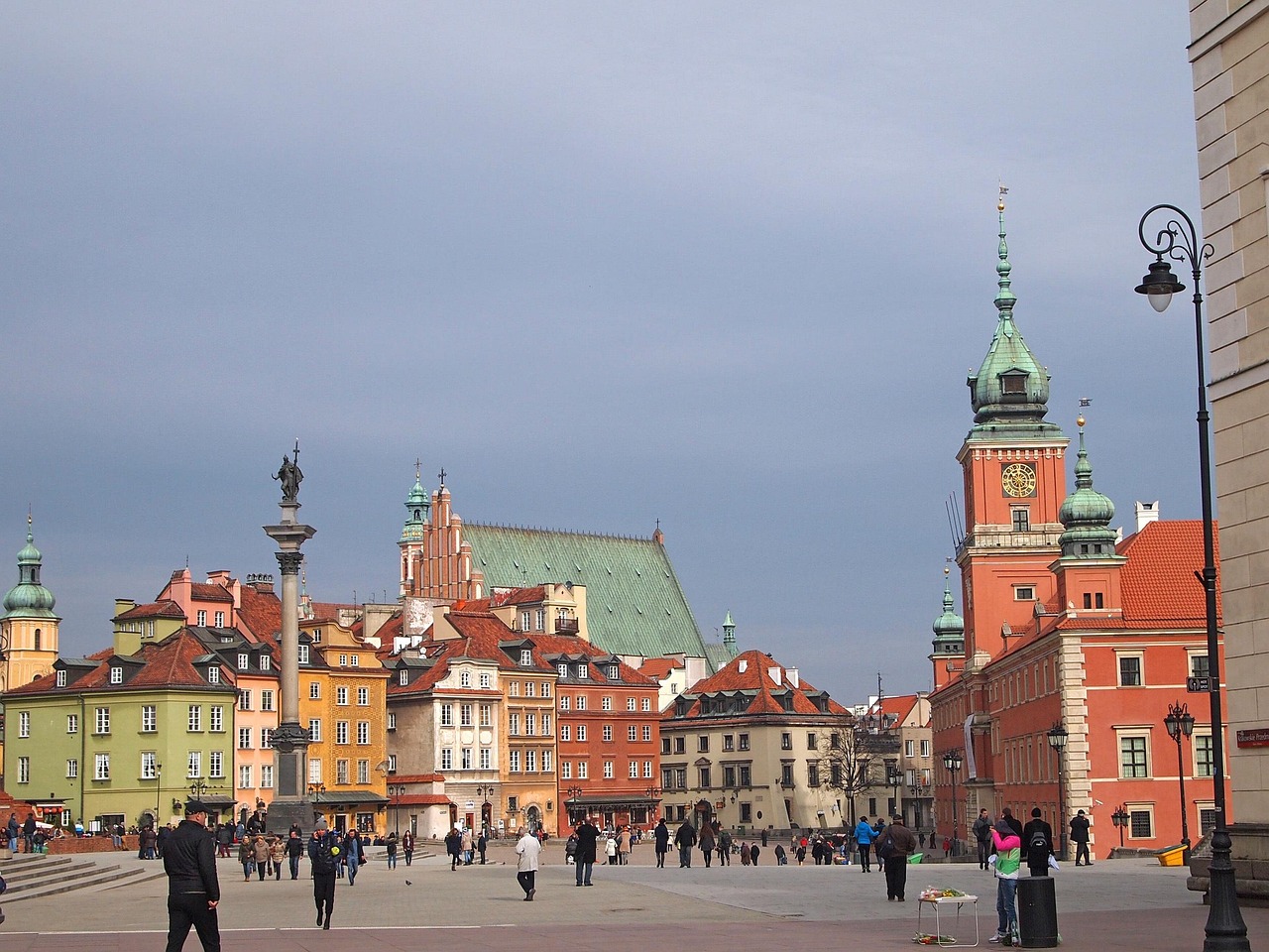 A bustling square in Warsaw, Poland, featuring colorful buildings, the Royal Castle, and Sigismund's Column.