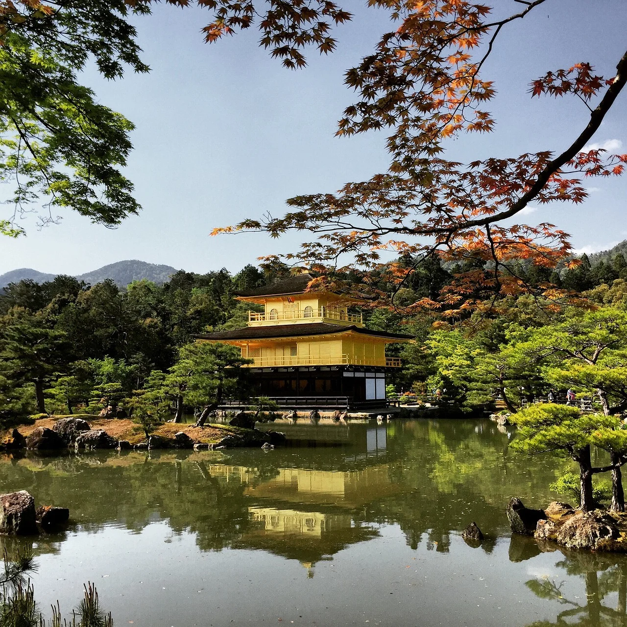 A serene view of the Kinkaku-ji temple, also known as the Golden Pavilion, reflected in a calm pond surrounded by lush greenery and autumn foliage.