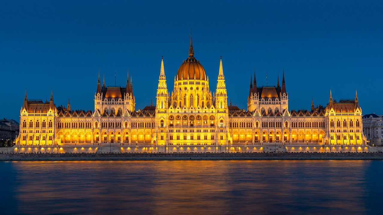 A grand, illuminated building with a large dome and multiple spires, reflected in calm water at night.